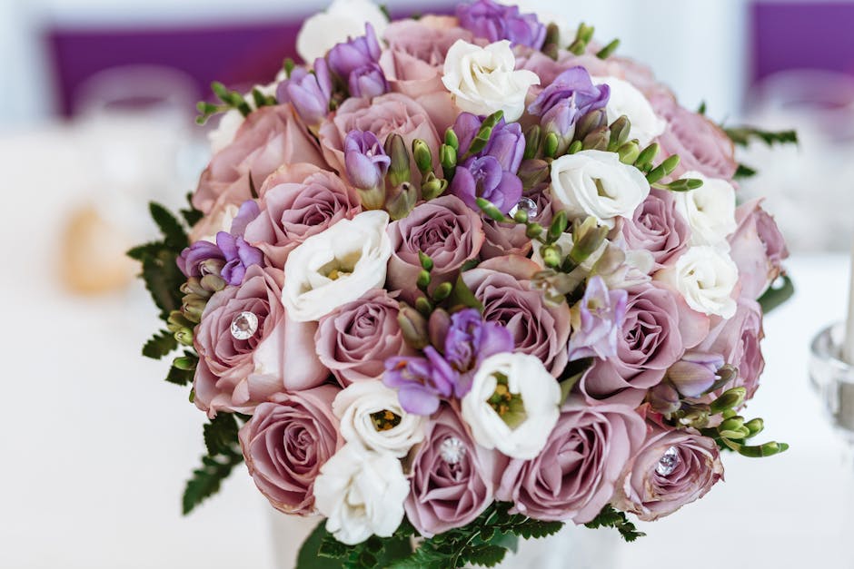 A close-up of a floral bouquet featuring soft pink roses, white eustoma, and purple lisianthus, arranged in a rounded, lush style. The bouquet includes deep green fern leaves interspersed among the fl