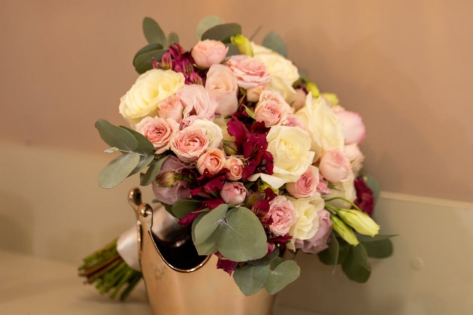 A bouquet of fresh flowers arranged in a rounded style, featuring pale pink roses, cream-colored roses, and deep burgundy blossoms, accented with silvery green eucalyptus leaves. The flowers are wrapp