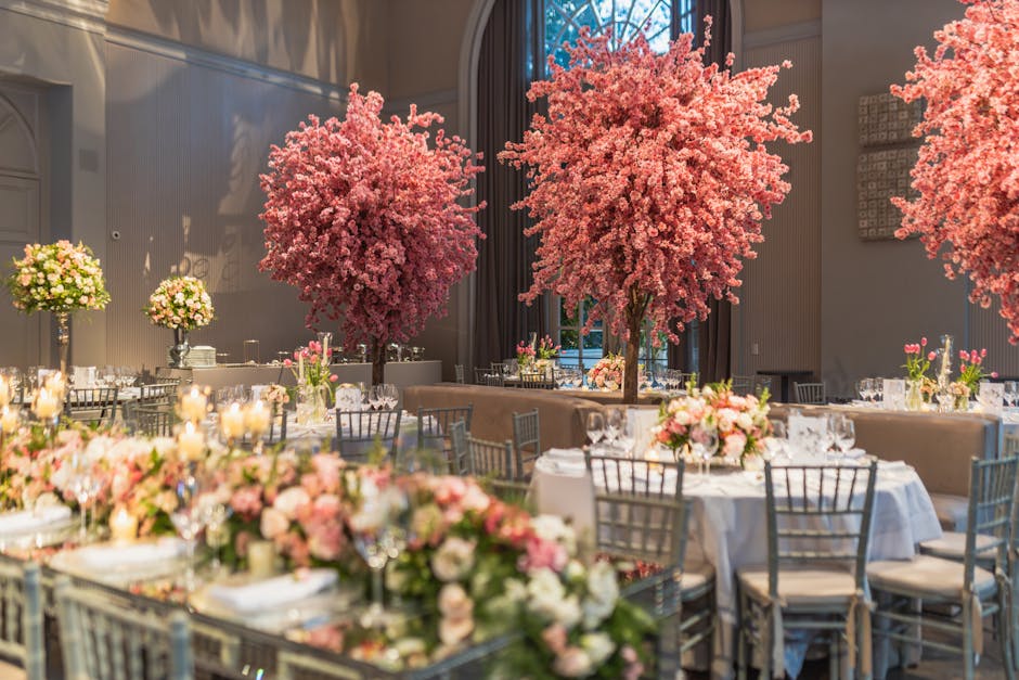 An elegant wedding reception setup featuring multiple large pink cherry blossom trees as central floral displays. Surrounding the trees are round tables draped in white tablecloths, decorated with arr