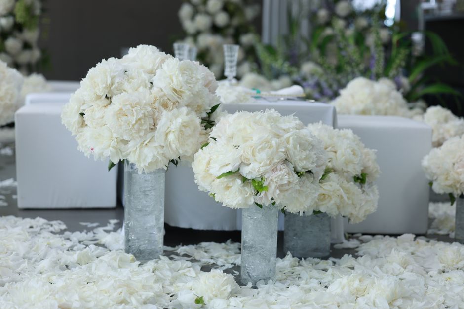 Two elegant floral arrangements featuring large, fluffy white peonies and roses arranged in tall, clear glass vases. The bouquets are set on a surface scattered with white flower petals, with addition