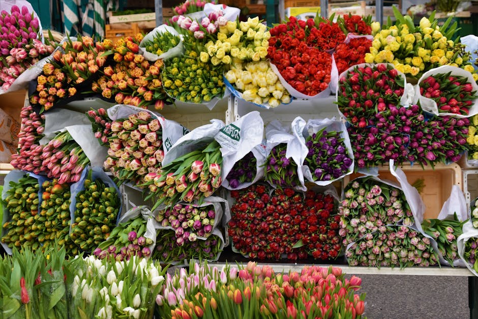 A vibrant display of flower bouquets arranged on a market stall, featuring a variety of tulips, roses, and other blooms in shades of pink, red, yellow, purple, and white. The flowers are wrapped in wh