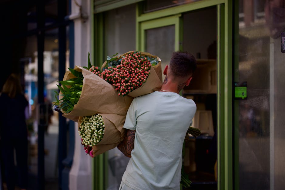 A person with a short haircut, wearing a light-colored t-shirt, is seen from behind carrying a large bouquet of fresh flowers wrapped in brown craft paper. The bouquet features an assortment of pink, 