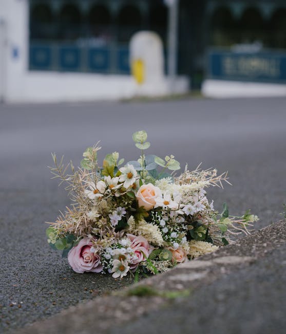 A delicate floral arrangement resting on an asphalt surface outdoors, consisting of soft pink roses, white daisies, and small filler flowers, accented with pale green foliage and wispy, dried botanica