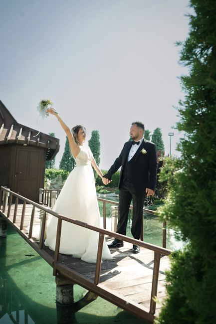 A bride and groom holding hands on a small wooden bridge outdoors, with the bride holding a cascading bouquet of white and green flowers aloft. The bride wears a white wedding gown with lace details, 