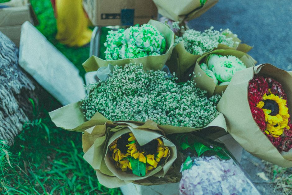 A collection of freshly arranged flower bouquets wrapped in light brown paper and placed outdoors on the ground, displaying vibrant colours including green, white, yellow, red, and purple. The bouquet