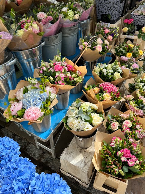 An array of freshly cut floral bouquets arranged on a blue table at a florist stall, featuring pink and white roses, blue hydrangeas, and a mix of greenery, all wrapped in brown kraft paper or placed 