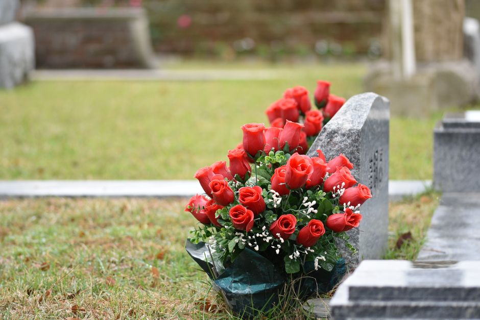 A bouquet arrangement of fresh, vibrant red roses with tightly closed buds and open petals, accompanied by small white filler flowers and lush green foliage. The bouquet is wrapped in dark green flora